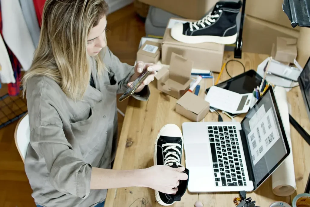 Perempuan memotret sepatu untuk dijual secara online di ruang kerja rumahan dengan laptop dan paket siap kirim
